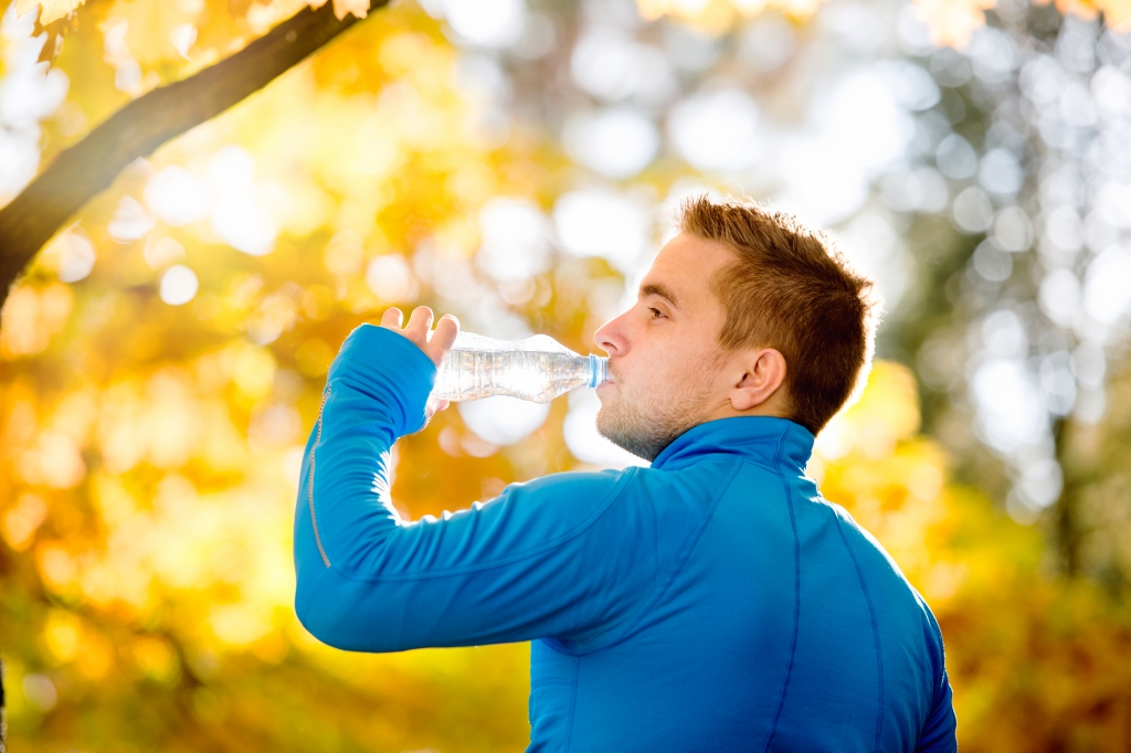 Young handsome hipster runner in blue sweatshirt in sunny autumn nature drinking water from bottle, rear view