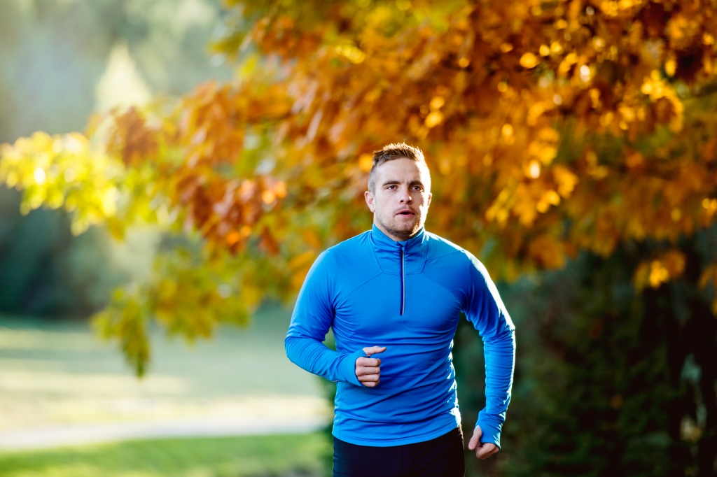 Young handsome hipster athlete in blue sweatshirt running outside in colorful sunny autumn nature